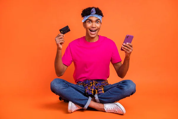 Foto de tamaño corporal de cuerpo entero del chico sorprendido que muestra la tarjeta bancaria manteniendo el teléfono inteligente aislado color naranja brillante fondo —  Fotos de Stock