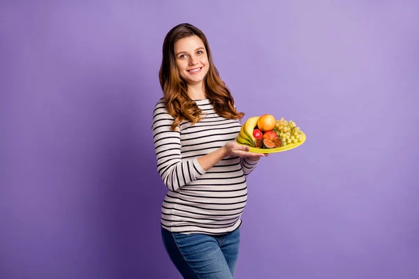 Foto de mulher doce adorável esperando criança usar barriga camisa listrada segurando prato de frutas isolado cor roxa fundo — Fotografia de Stock