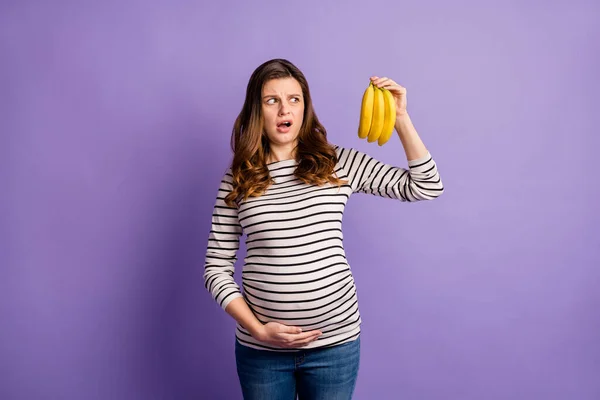 Foto retrato de una mujer embarazada molesta sosteniendo mirando plátanos aislados sobre fondo violeta vivo — Foto de Stock