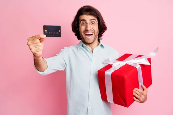 Portret van jonge knappe gelukkig dolblij man man man tonen credit card houden rode doos gift wear bril geïsoleerd op roze kleur achtergrond — Stockfoto