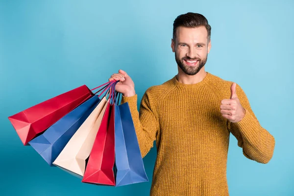 Foto de joven guapo sonriente positivo alegre hombre mantenga bolsas de compras muestran pulgar hacia arriba aislado sobre fondo de color azul — Foto de Stock