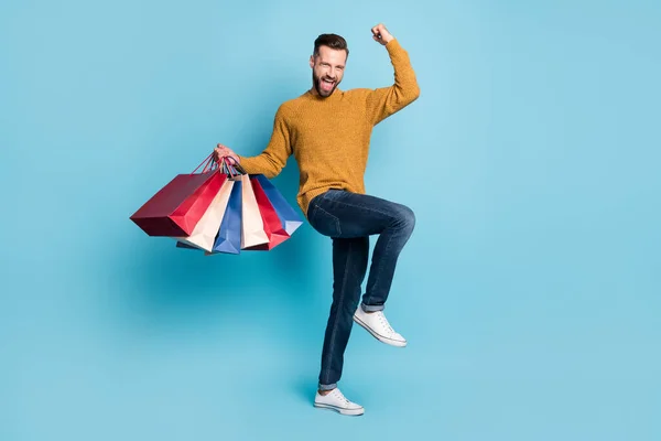 Zdjęcie młodego wesołego podekscytowany szalony pozytywny człowiek trzymać torby na zakupy podnieść pięść w zwycięstwie odizolowany na tle niebieskiego koloru — Zdjęcie stockowe