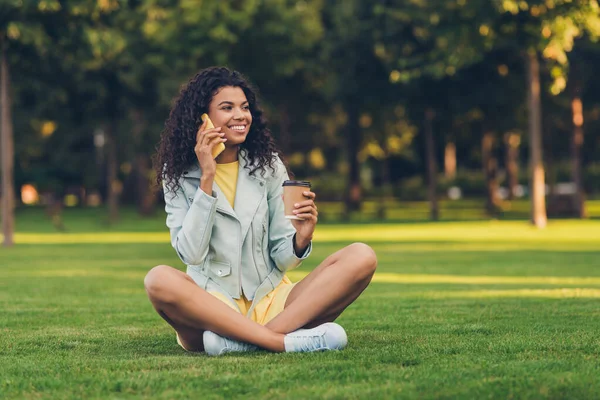 Retrato de bela menina alegre na moda sentado na grama bebendo bebida chamando amigo passatempo ao ar livre — Fotografia de Stock