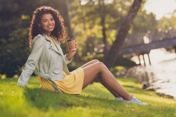 Foto de senhora sentar grama segurar copo takeout café olhar câmera usar calçado vestido amarelo no parque verde ao ar livre — Fotografia de Stock
