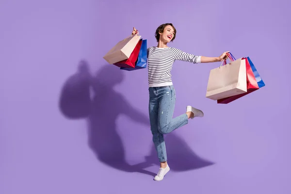 Foto de comprimento total de mulher comprador doce usar roupas casuais dançando segurando sacos de compras coloridos isolado fundo cor violeta — Fotografia de Stock