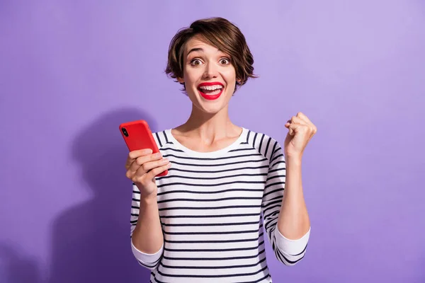 Фото збудженої красивої дівчини тримайте телефон підняти кулак в смугастій сорочці ізольований фіолетовий кольоровий фон — стокове фото