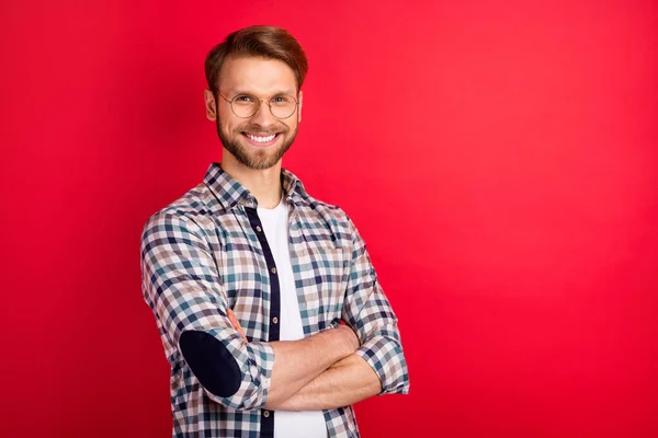 Fénykép jóképű aranyos fiatal srác viselni kockás ing szemüveg mosolygó karok keresztbe üres tér elszigetelt piros háttér — Stock Fotó
