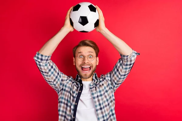 Fotografie vzrušený funky mladý chlap nosit kostkované košile brýle házení nohy míč s úsměvem izolované červené barvy pozadí — Stock fotografie