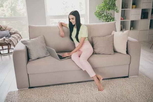 Zdjęcie młodej uroczej pani siedzieć kanapa trzymać netbook promieni uśmiech nosić zielony t-shirt różowe spodnie boso w pomieszczeniach — Zdjęcie stockowe