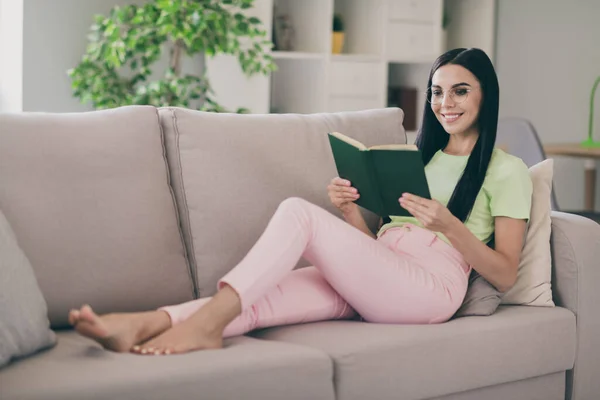 Φωτογραφία από χαρούμενα clam νεαρή κοπέλα θέσει καναπέ μαξιλάρια κρατήστε το βιβλίο διαβάσει ξυπόλυτος φορούν γυαλιά πράσινο t-shirt ροζ παντελόνι σε εσωτερικούς χώρους — Φωτογραφία Αρχείου