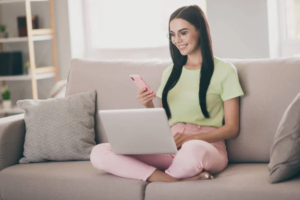 Foto de niña alegre sentarse sofá piernas cruzadas sostener netbook teléfono llevar gafas verde camiseta pantalones de color rosa en el interior — Foto de Stock