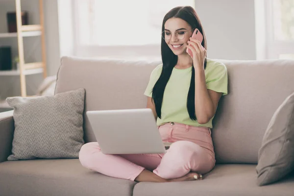 Фото очаровательной девушки сидеть диван держать телефон разговор ноутбук смотреть экран носить очки зеленые футболки розовые брюки в помещении — стоковое фото