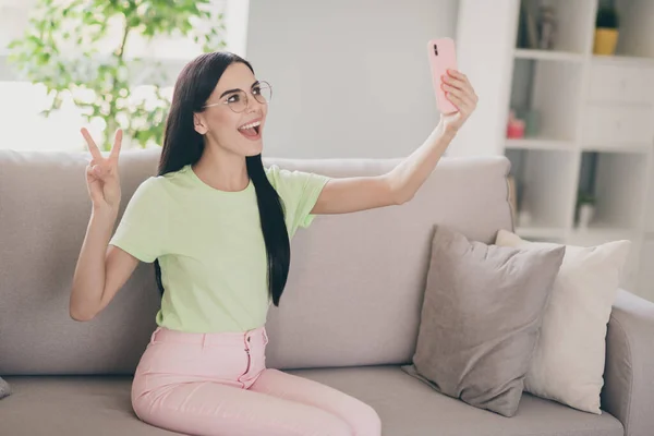 Foto van grappig meisje zit bank hold telefoon nemen selfie show v-teken gek gezicht dragen bril groen t-shirt roze broek binnen — Stockfoto