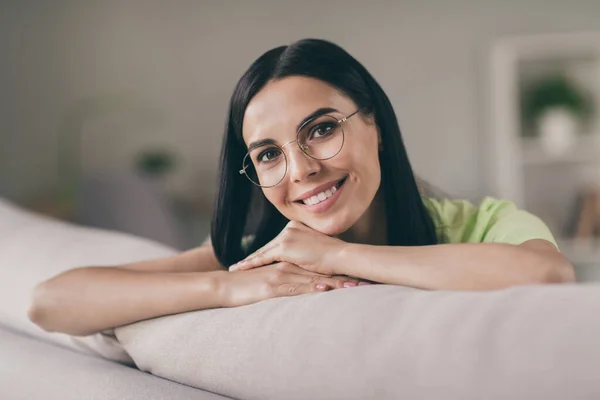 Foto van dromerige jonge dame zitten sofa armen kin witte glimlach kijken camera dragen bril groen t-shirt binnen — Stockfoto