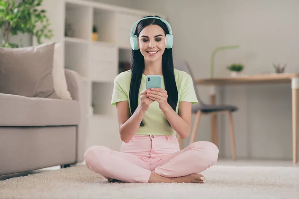 Foto de linda chica joven sentarse piso celebrar el aspecto del teléfono celular pantalla sonrisa blanca usar auriculares camiseta verde pantalones de color rosa en el interior — Foto de Stock