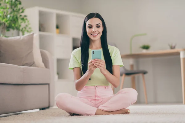 Fotografie veselé mírumilovné dívka sedět podlaha zkřížené nohy držet telefon lesklý úsměv vzhled obrazovky nosit zelené tričko růžové kalhoty uvnitř — Stock fotografie