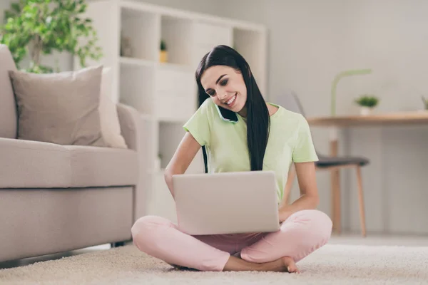 Retrato de una encantadora chica alegre enfocada sentada en posición de loto usando computadora portátil hablando en el teléfono en el plano interior ligero — Foto de Stock