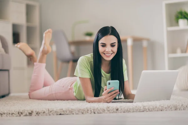 Retrato de una chica de pelo largo bastante alegre enfocada acostada en la alfombra usando un dispositivo portátil blogueando en el plano interior ligero — Foto de Stock