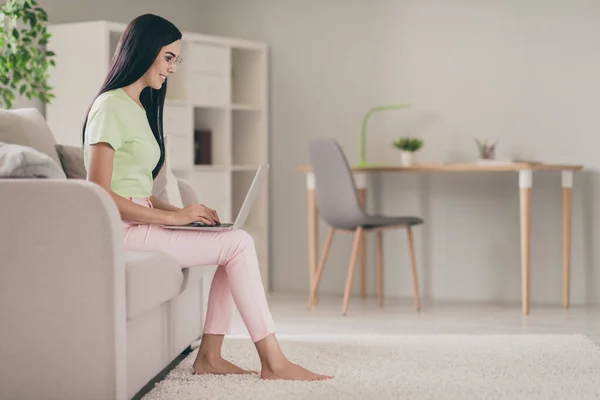 Profile side view of pretty wesoły skupiony długowłosy dziewczyna siedzi na kanapie za pomocą laptopa pisanie e-mail w jasnym pokoju — Zdjęcie stockowe