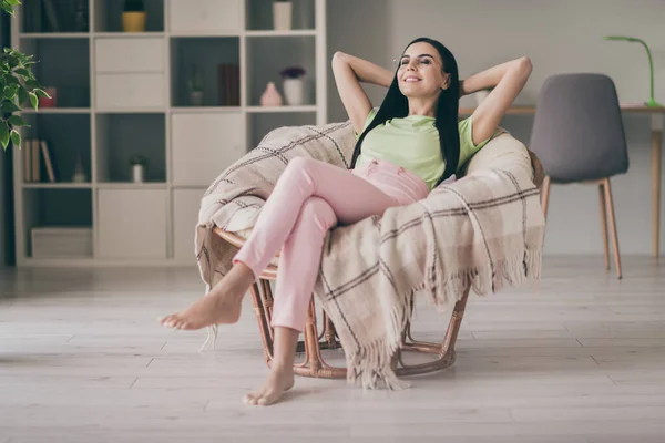 Retrato de chica alegre bastante soñadora sentada en silla acogedora descalza sobre un velo a cuadros disfrutando del descanso en la habitación interior de luz — Foto de Stock