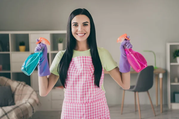 Retrato de empregada de cabelos compridos muito alegre segurando em mãos garrafas pulverizadores limpeza na luz interior sala de estar — Fotografia de Stock