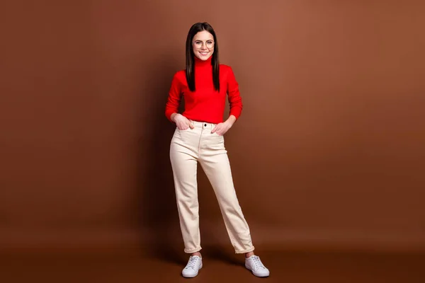 Foto wanita muda yang cantik menawan mengenakan sweater merah berdiri saku lengan tersenyum terisolasi warna latar belakang coklat — Stok Foto