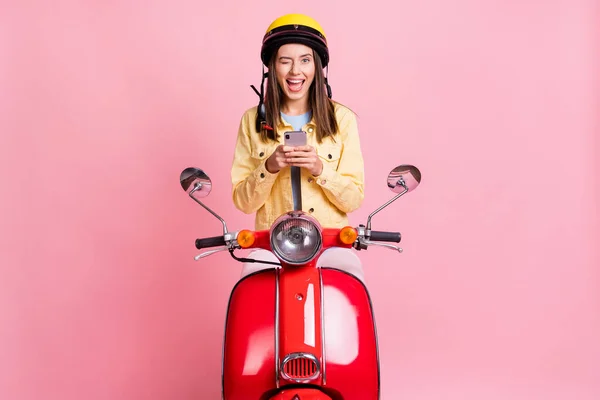 Foto de jovem positivo bela garota piscadela usar telefone usar capacete amarelo passeio moto vermelha isolada no fundo cor-de-rosa — Fotografia de Stock