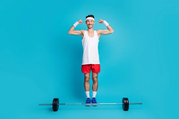 Full length φωτογραφία του νεαρού άνδρα ευτυχής θετικό χαμόγελο κρατήστε τα χέρια δείχνουν μυς barbell bodybuilding απομονώνονται σε μπλε χρώμα φόντο — Φωτογραφία Αρχείου