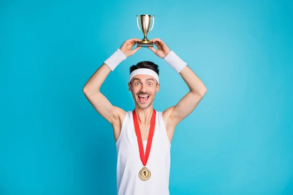 Foto de morena sorprendida hombre feliz usar diadema medalla de oro levantar trofeo por encima de la cabeza ganar aislado sobre fondo de color azul —  Fotos de Stock