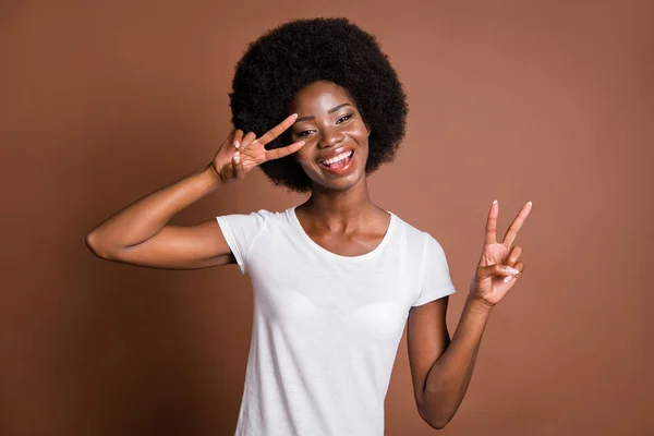 Foto porträtt av flicka som visar v-tecken gest båda händerna i vit outfit leende isolerad på brun färg bakgrund — Stockfoto