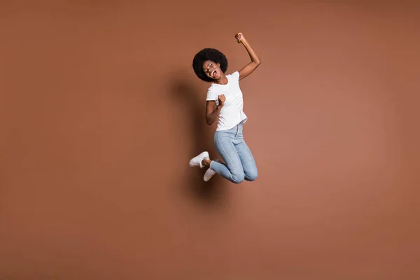 Фото энергичной девушки прыгать с открытым ртом поднять кулаки носить белые футболки джинсы обувь изолированный коричневый цвет фона — стоковое фото