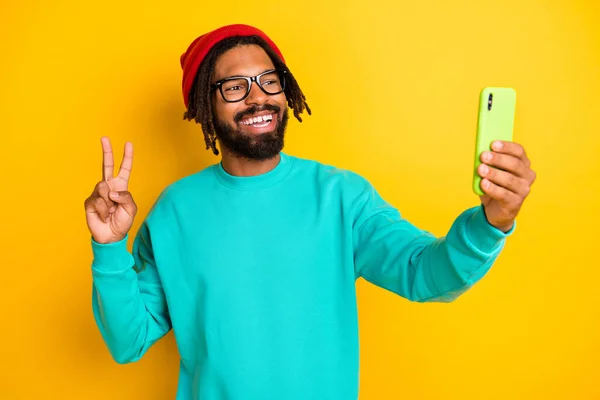 Portret van brunette optimistische man doen selfie show v-teken slijtage cap bril blauw pullover geïsoleerd op geel levendige kleur achtergrond — Stockfoto