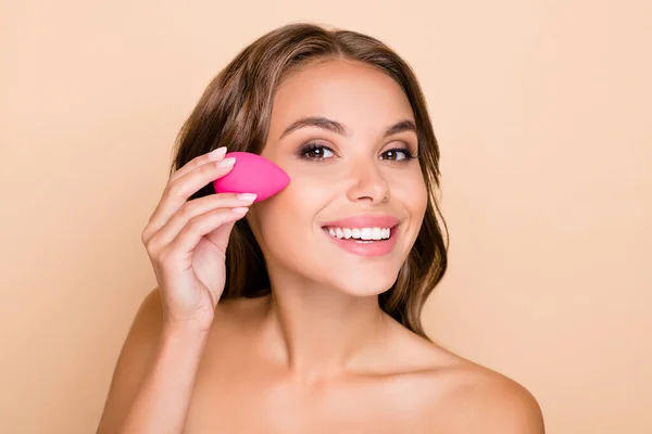 Foto de una joven hermosa mujer sonriente aplicando una base en la mejilla con una licuadora de belleza aislada en un fondo de color beige — Foto de Stock
