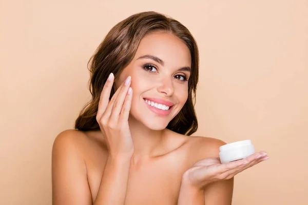 Foto di giovane splendida ragazza felice senza vestiti applicare crema idratante sulle guance isolate su sfondo di colore beige — Foto Stock