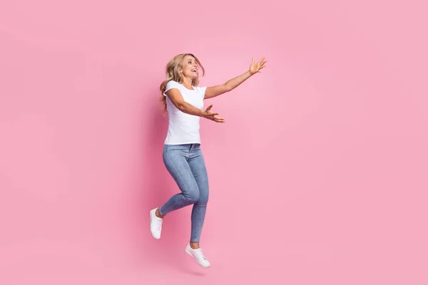 女性の写真ジャンプストレッチ手キャッチ見て空のスペースは白いTシャツのジーンズの靴を着用ピンク色の背景を隔離 — ストック写真
