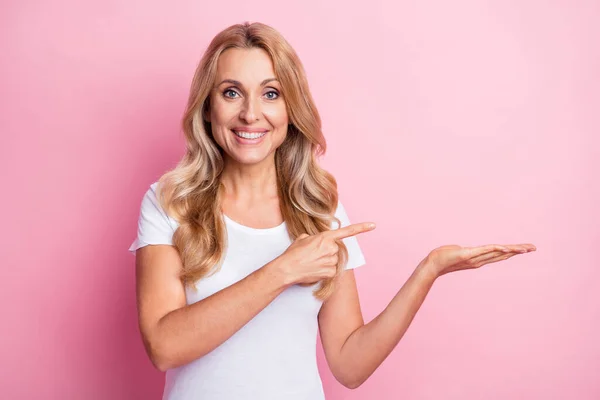 Foto av söt optimistisk blond dam håll punkt tomt utrymme slitage vit t-shirt isolerad på pastell rosa färg bakgrund — Stockfoto