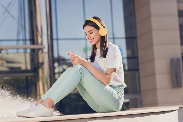 Full size profile photo of optimistic brunette lady sit with headphones wear white cloth trousers sneakers walking in park