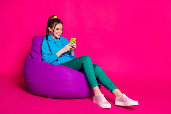 全长照片，笑容满面的女人身穿蓝色连帽衫，躺在紫色豆袋里，拿着现代装置隔离的粉色底色 — 图库照片