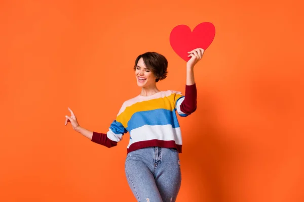 Foto de mulher doce adorável usar camisola listrada dança segurando coração vermelho apontando espaço vazio isolado cor de fundo laranja — Fotografia de Stock