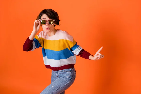 Fotografie okouzlující legrační žena nosit pruhované svetr tanec paže tmavé brýle ukazující prázdný prostor izolované oranžové barvy pozadí — Stock fotografie