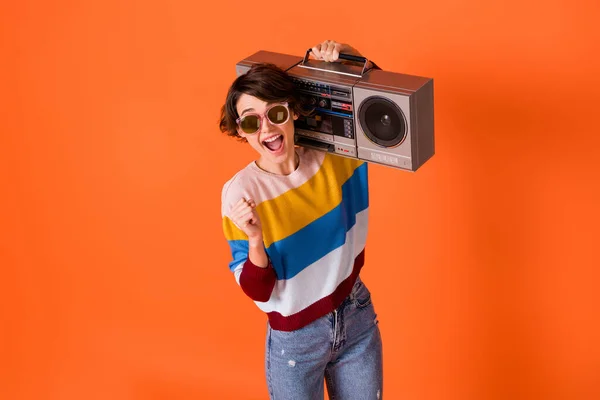 Foto di funky fortunato giovane donna indossare maglione a righe occhiali alzando pugno che tiene boombox isolato colore arancione sfondo — Foto Stock