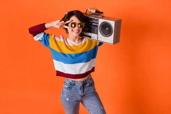 Ritratto di divertente allegra ragazza portare boom box dita mostrano saluti v-segno vicino occhio isolato su sfondo di colore arancione — Foto Stock