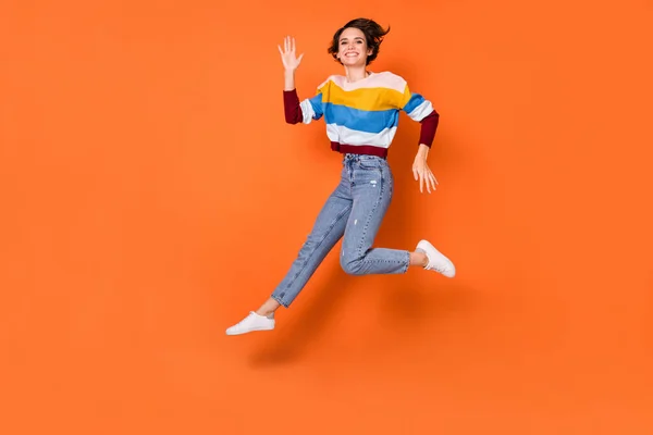 Photo of funky adorable young lady dressed striped pullover jumping waving arm isolated orange color background — Stock Photo, Image