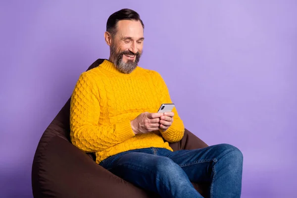 Foto de hombre sit beanbag hold phone look screen wear yellow sweater jeans isolated purple background —  Fotos de Stock