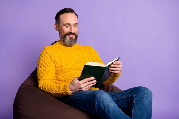 Portret van een vrolijke man zitten zachte stoel handen houden leesboek tandenglimlach geïsoleerd op paarse kleur achtergrond — Stockfoto