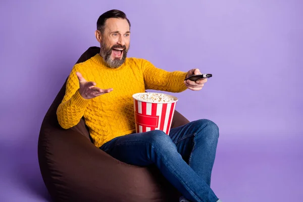 Фото человека сидеть бобовый мешок держать пульт дистанционного управления коробку попкорн носить желтый свитер джинсы изолированный фиолетовый фон — стоковое фото