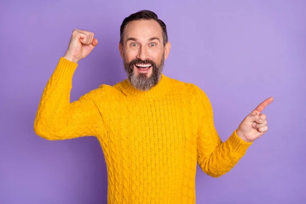 Фото веселого мужчины с поднятием кулака указывает на пустое пространство указательного пальца носить желтый свитер изолированный фиолетовый фон — стоковое фото