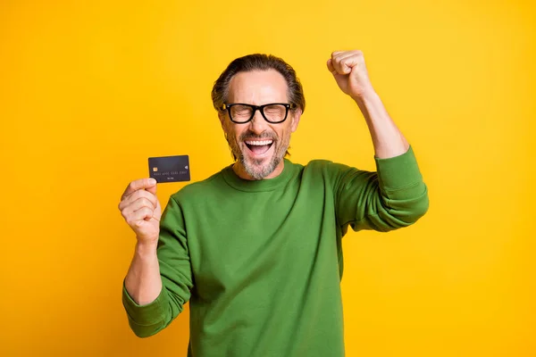 Foto van opgewonden man presenteren plastic debetkaart verhogen vuist slijtage bril groen trui geïsoleerde gele kleur achtergrond — Stockfoto