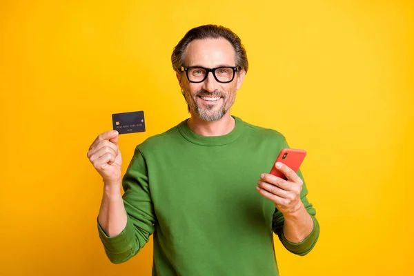 Foto de hombre alegre demostrar teléfono móvil tarjeta de débito gafas de desgaste jersey verde aislado color amarillo fondo — Foto de Stock