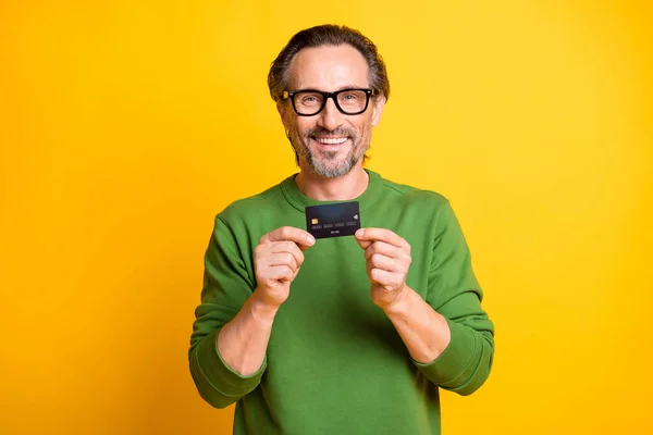 Retrato fotográfico del hombre con gafas que muestran la tarjeta bancaria de débito sonriendo en ropa verde aislada sobre fondo de color amarillo brillante — Foto de Stock